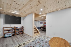 A room with a wooden ceiling, a flat-screen TV mounted on the wall, a bookshelf, a floral-patterned rug, and a wooden staircase leading to another room. - Gaylord Lodge:  Forest Escape: Golf, Ski, Fish and Sleep 8! (Gaylord)