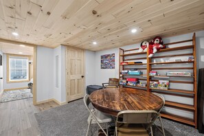 Ein Zimmer mit Holzbalkendecke, einem runden Tisch mit Stühlen, einem Bücherregal mit Gesellschaftsspielen und einer Tür.