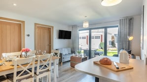 Eine moderne Küche mit Kochinsel, Essbereich mit weißen Stühlen, einem flachbildfernseher an der Wand und einer großen Schiebeglastür, die auf eine Außenpatio führt.