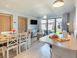 Eine moderne Küche mit Kochinsel, Essbereich mit weißen Stühlen, einem flachbildfernseher an der Wand und einer großen Schiebeglastür, die auf eine Außenpatio führt.