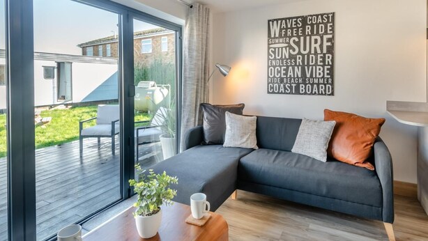 Ein modernes Wohnzimmer mit großem Fenster, einem grauen Sofa, einem Topf mit Pflanze und einer Wand mit motivierenden Sprüchen.