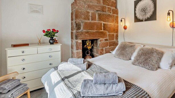 Un dormitorio con chimenea de piedra, una cama con ropa de cama gris y blanca, un tocador blanco y una lámpara fijada a la pared.
