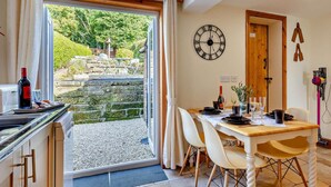 Un comedor con una mesa dispuesta para dos personas, un reloj en la pared y vistas a un jardín a través de un ventanal.