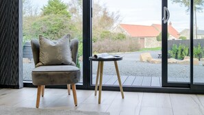 A modern room with a grey armchair, a small table, and a view of a rural landscape through large glass doors. - 3 Bed in Belford (oc-78058) (Easington)