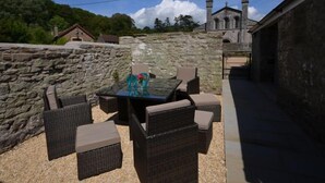 Eine Terrasse im Freien mit Korbmöbeln, einer Steinmauer und einem Gebäude mit einem Glockenturm im Hintergrund.