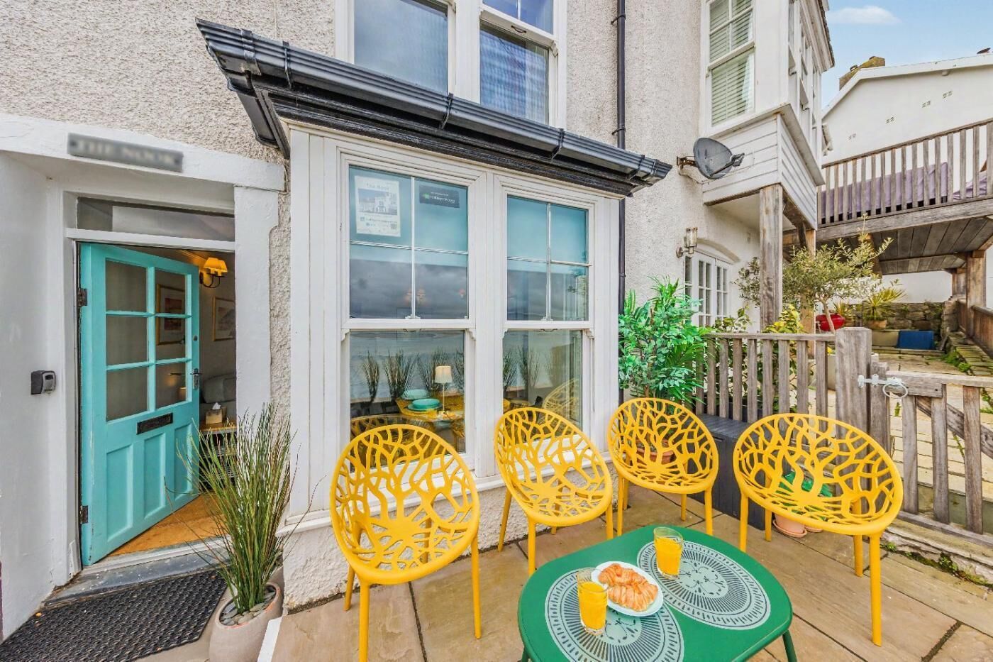A patio with yellow chairs and a table, a turquoise door, and a white building with a window.