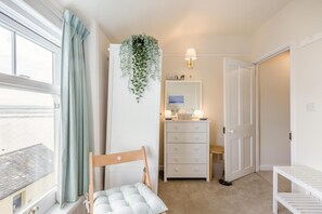 Ein Schlafzimmer mit Fenster, einer hängenden Pflanze, einer Kommode mit Schubladen, einem Nachttisch und einem Stuhl.