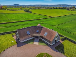 Une grande maison moderne, dotée d’un toit en pente, entourée de vastes champs verdoyants et surplombant un ciel dégagé.