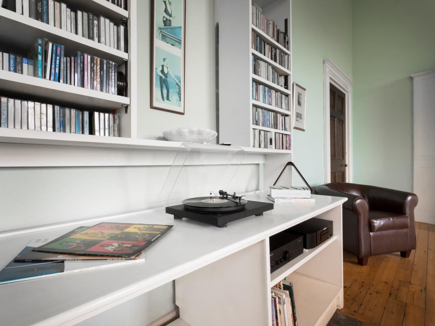 Una habitación con un escritorio blanco, estanterías, un sillón de cuero y un tocadiscos.