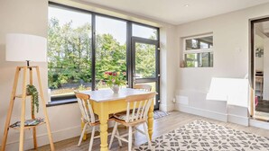 Ein modernes Zimmer mit Esstisch, Stühlen, einer Stehlampe und großen Fenstern mit Blick auf Grünflächen.