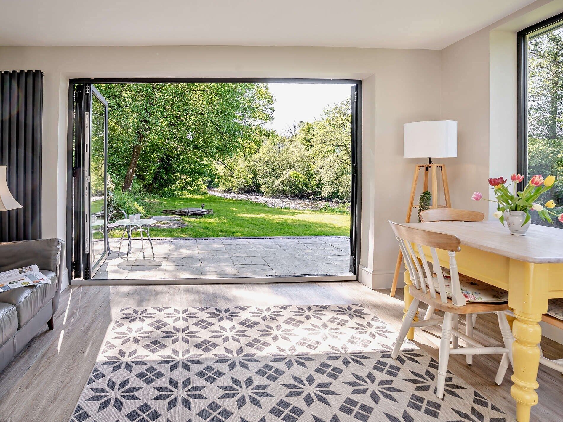 A modern interior with a sliding glass door leading to a garden, a dining area with a table and chairs, and a sofa.