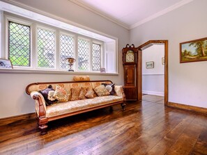 A room with a wooden floor, a vintage sofa with patterned cushions, a tall grandfather clock, and a window with a view of greenery.