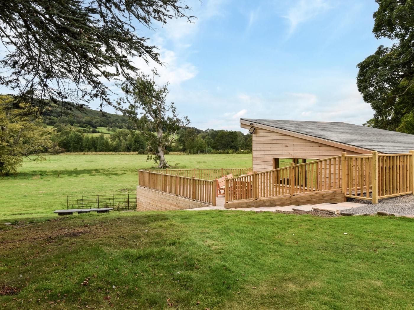 A wooden cabin with a porch and a staircase leading to a deck, surrounded by a grassy field and trees.