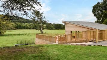 A wooden cabin with a porch and a staircase leading to a deck, surrounded by a grassy field and trees.