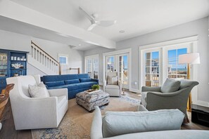 A spacious living room with a blue sofa, white armchairs, a patterned ottoman, and a ceiling fan. - Shoreview Manor | 35 ft to the beach | Private Pool and Hot Tub (Kitty Hawk)