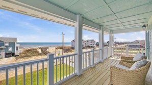 Eine überdachte Veranda mit einem Korbsessel, Blick auf den Strand und Häuser im Hintergrund.