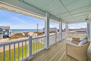 Eine überdachte Veranda mit einem Korbsessel, Blick auf den Strand und Häuser im Hintergrund.