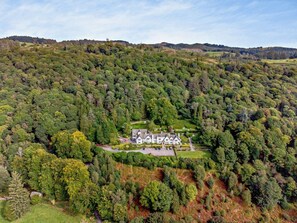 A large, multi-story house surrounded by dense forest, with a well-maintained lawn and driveway. - 2 Bed in Hawkshead (oc-llh12) (Claife)