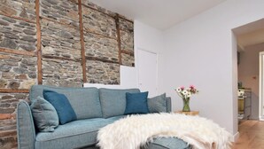 Ein modernes Wohnzimmer mit Steinwand, einem grauen Sofa mit blauen Kissen, einer flauschigen weißen Decke und einer Vase mit Blumen auf einem Beistelltisch.