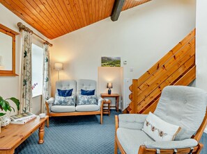 Ein gemütliches Zimmer mit Holzbalkendecke, einer Treppe, zwei Sesseln mit gemusterten Kissen, einem kleinen Tisch mit Lampe sowie einem Bücherregal.