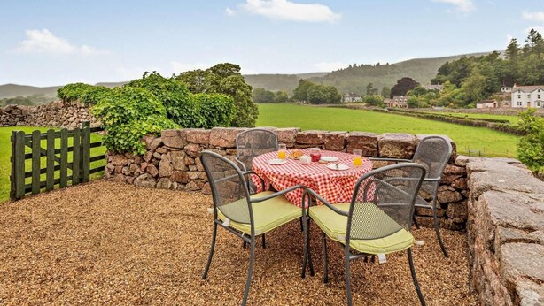Une terrasse avec une table dressée pour deux personnes, des murs en pierre et une vue sur un champ verdoyant et des maisons au loin.