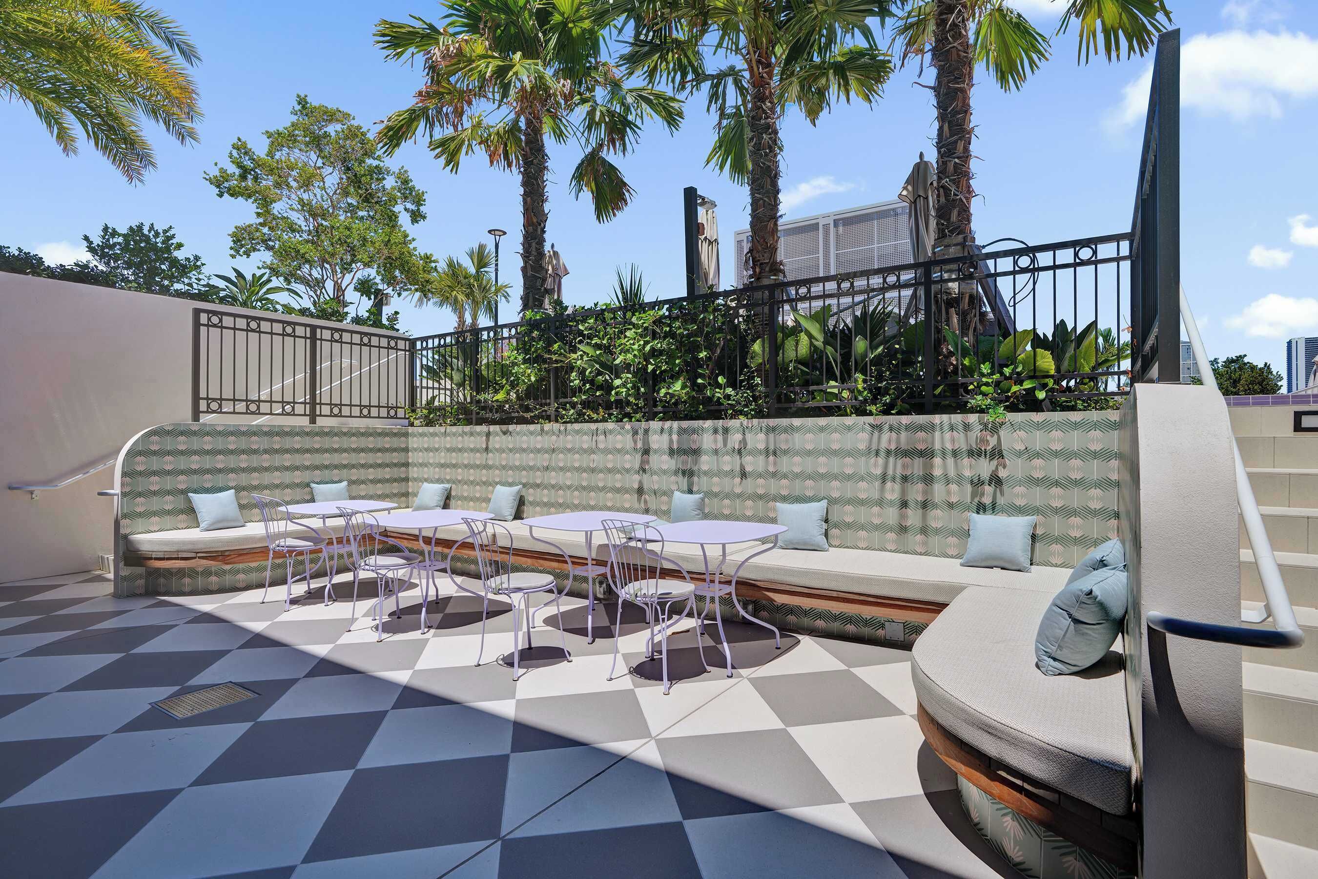 Un patio con piso a cuadros, una zona de estar con sofá y mesas, y vistas a palmeras y edificios.