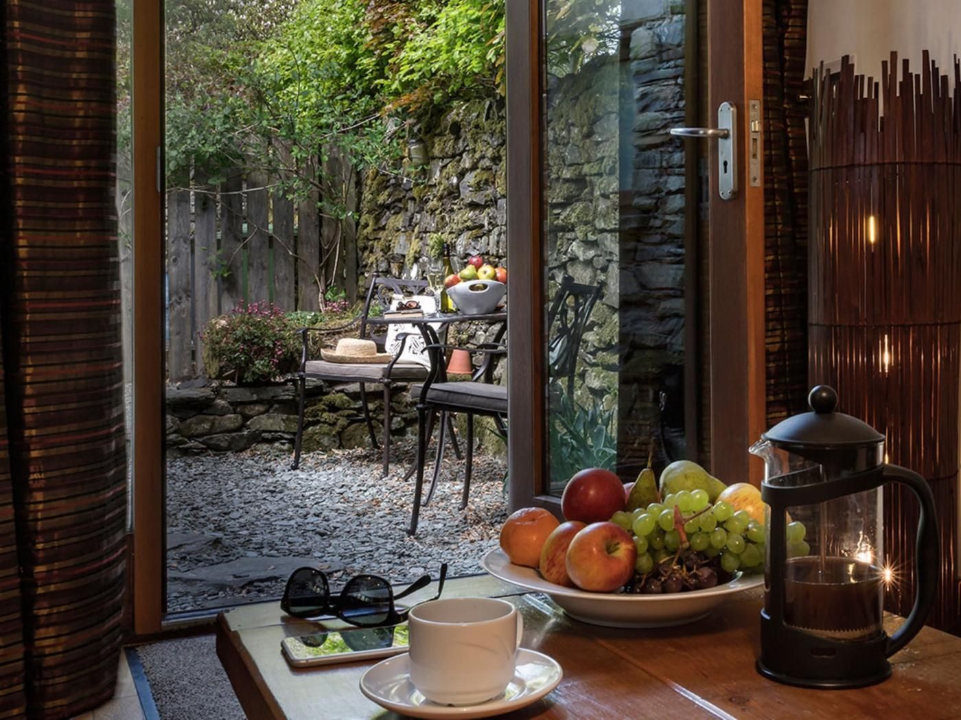 A cozy outdoor seating area with a stone wall, a table with a coffee pot, and a plate of fruit.