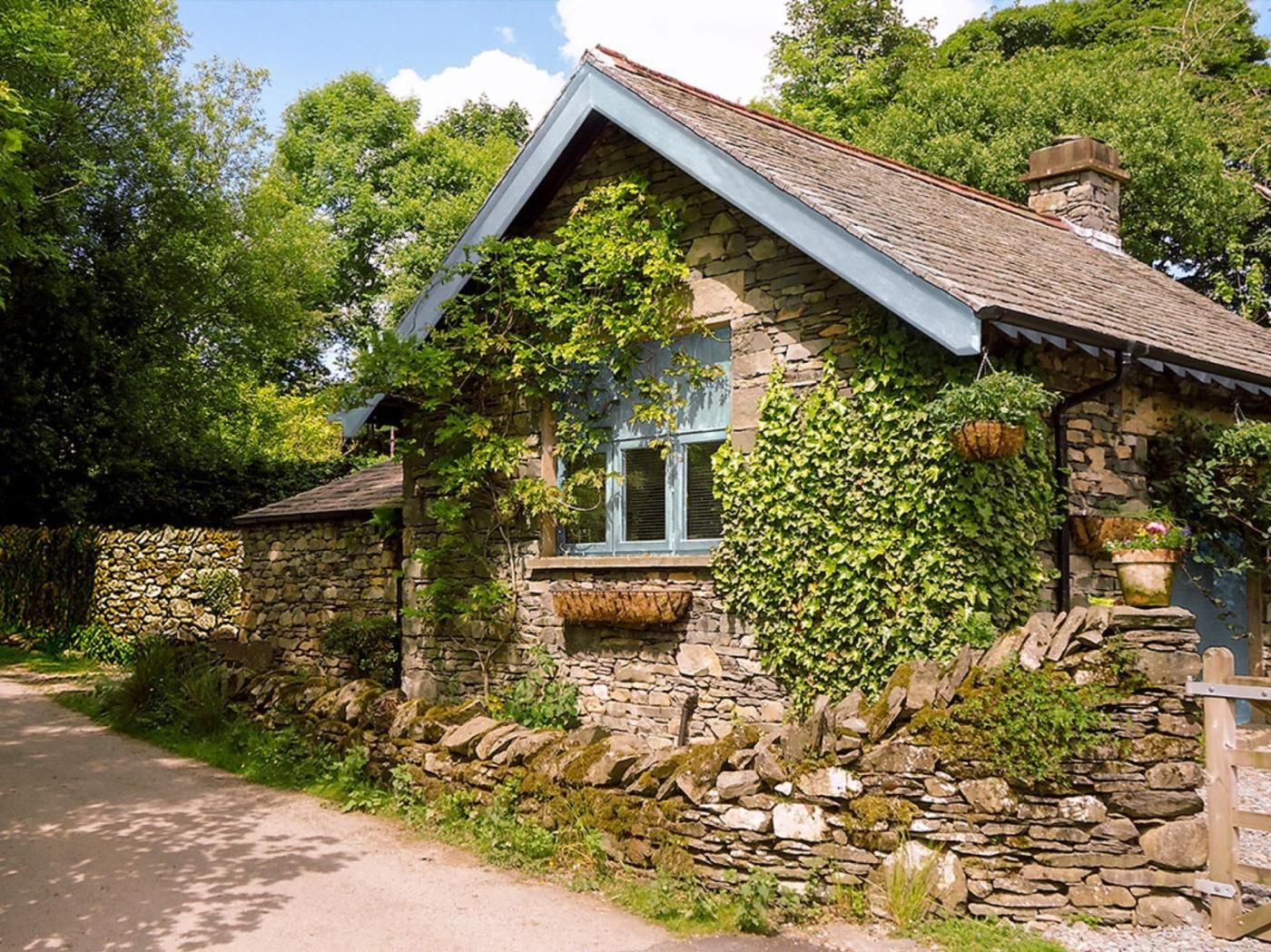 A stone cottage with a gabled roof, a window with shutters, and a stone wall with vegetation.