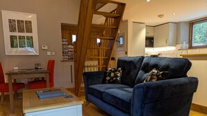Un salon avec un canapé bleu, une table basse en bois, un fauteuil rouge, un escalier en bois et, au fond, une cuisine.
