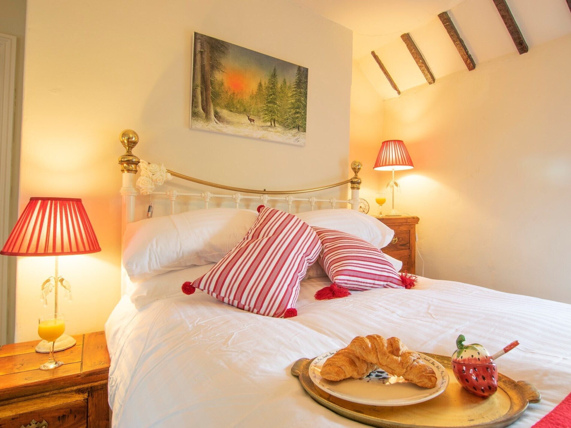 Una cama con ropa de cama blanca y almohadas a rayas rojas, un cuadro de un paisaje forestal y una bandeja con croissants y una taza con forma de fresa.