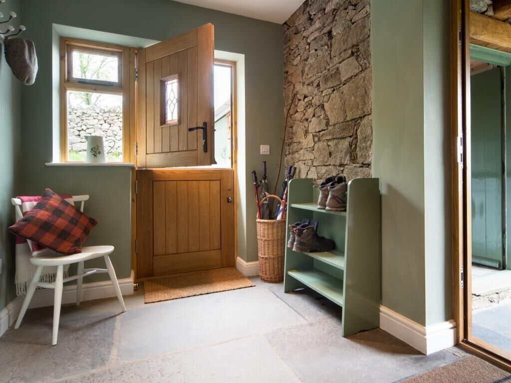 Una habitación con pared de piedra, puerta de madera, una silla, estante con zapatos y ventana con un florero.