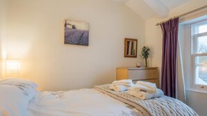 Een slaapkamer met een bed, nachtkastje, lamp, schilderij, raam met gordijnen en uitzicht.