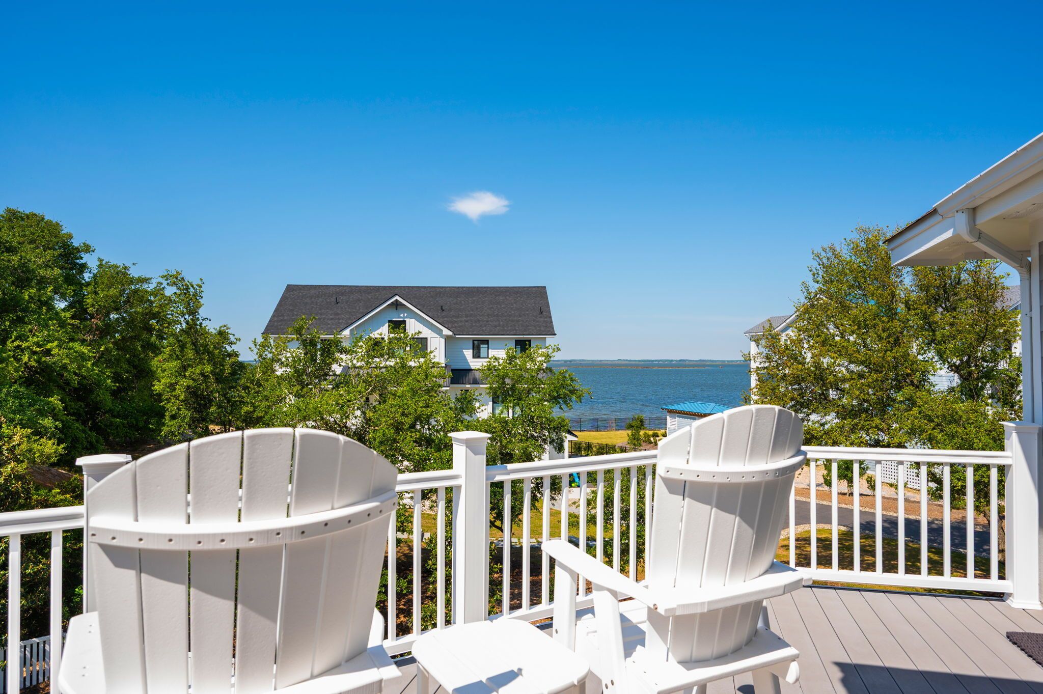 A deck with white Adirondack chairs overlooking a beach and a house.