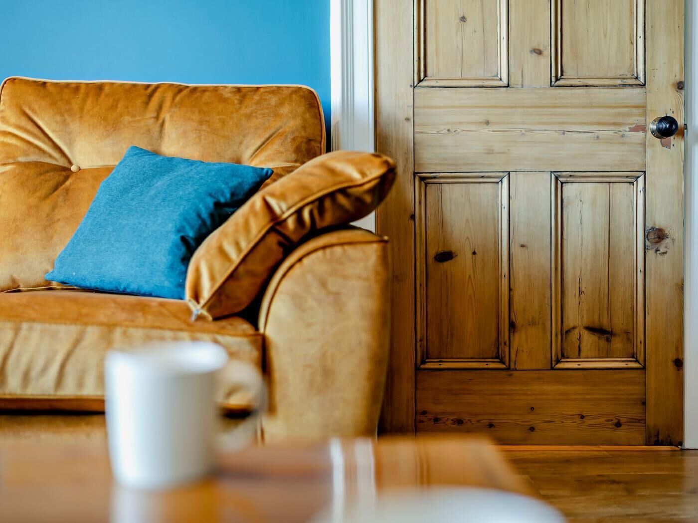 Een kamer met een houten deur, een bruine leren bank met een blauw kussen, en een witte mok op een tafel.