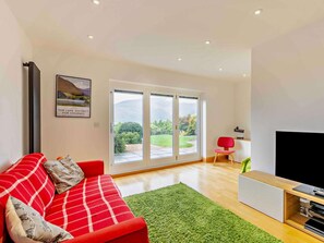 A modern living room with a red sofa, a green rug, a flat-screen TV, and a large window with a view of greenery. - 3 Bed in Underskiddaw (oc-sz377) (Underskiddaw)
