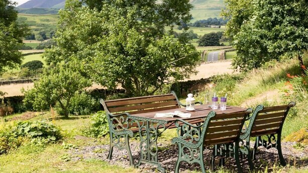 A wooden bench with metal armrests and a table with a lantern, positioned in a lush green landscape with hills in the background. - 2 Bed in Keswick (oc-sz330) (Saint John's Castlerigg and Wythburn)