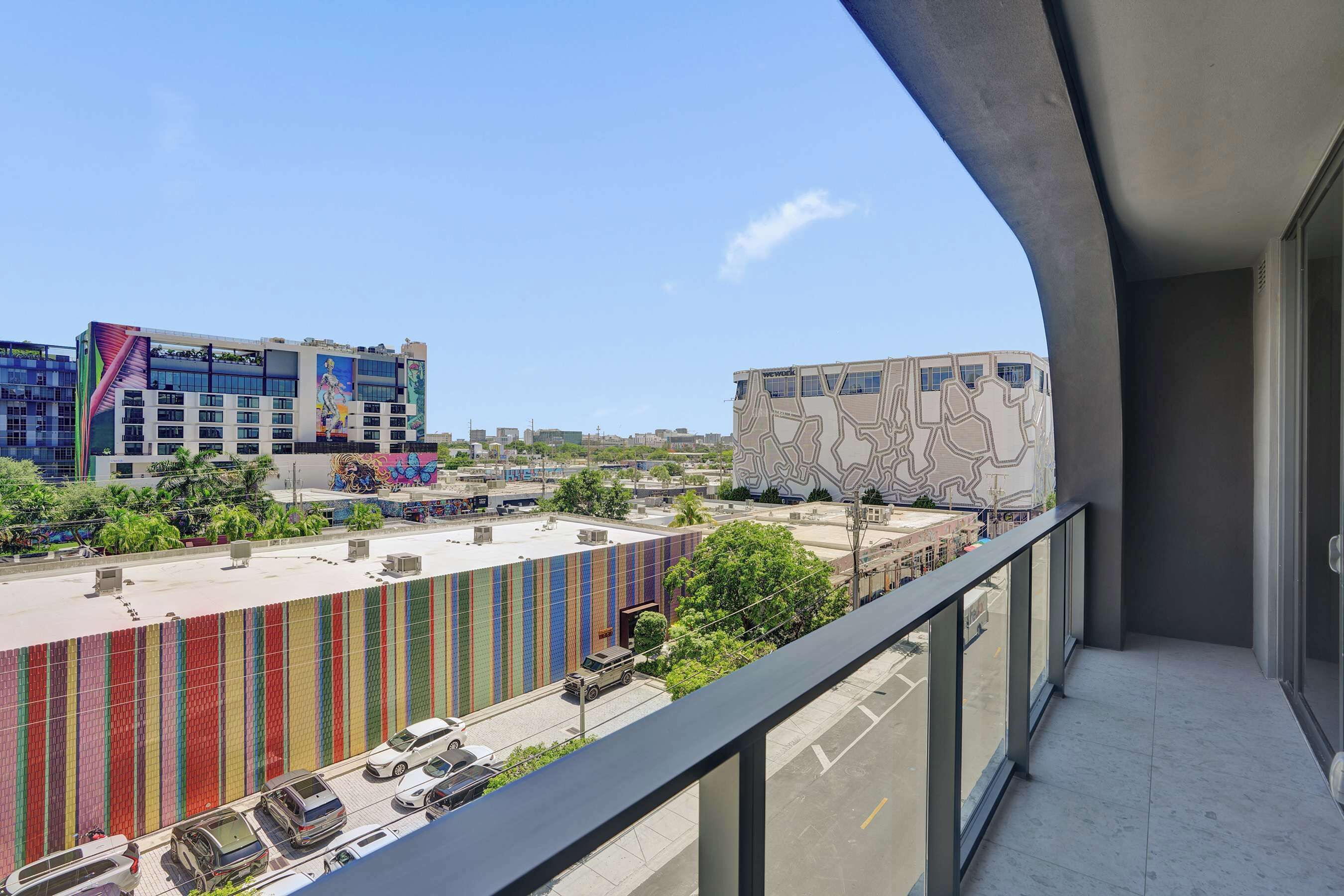 Uma varanda com vista para um edifício moderno, fachada colorida e um mural, um estacionamento e uma rua com carros.