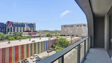 Uma varanda com vista para um edifício moderno, fachada colorida e um mural, um estacionamento e uma rua com carros.
