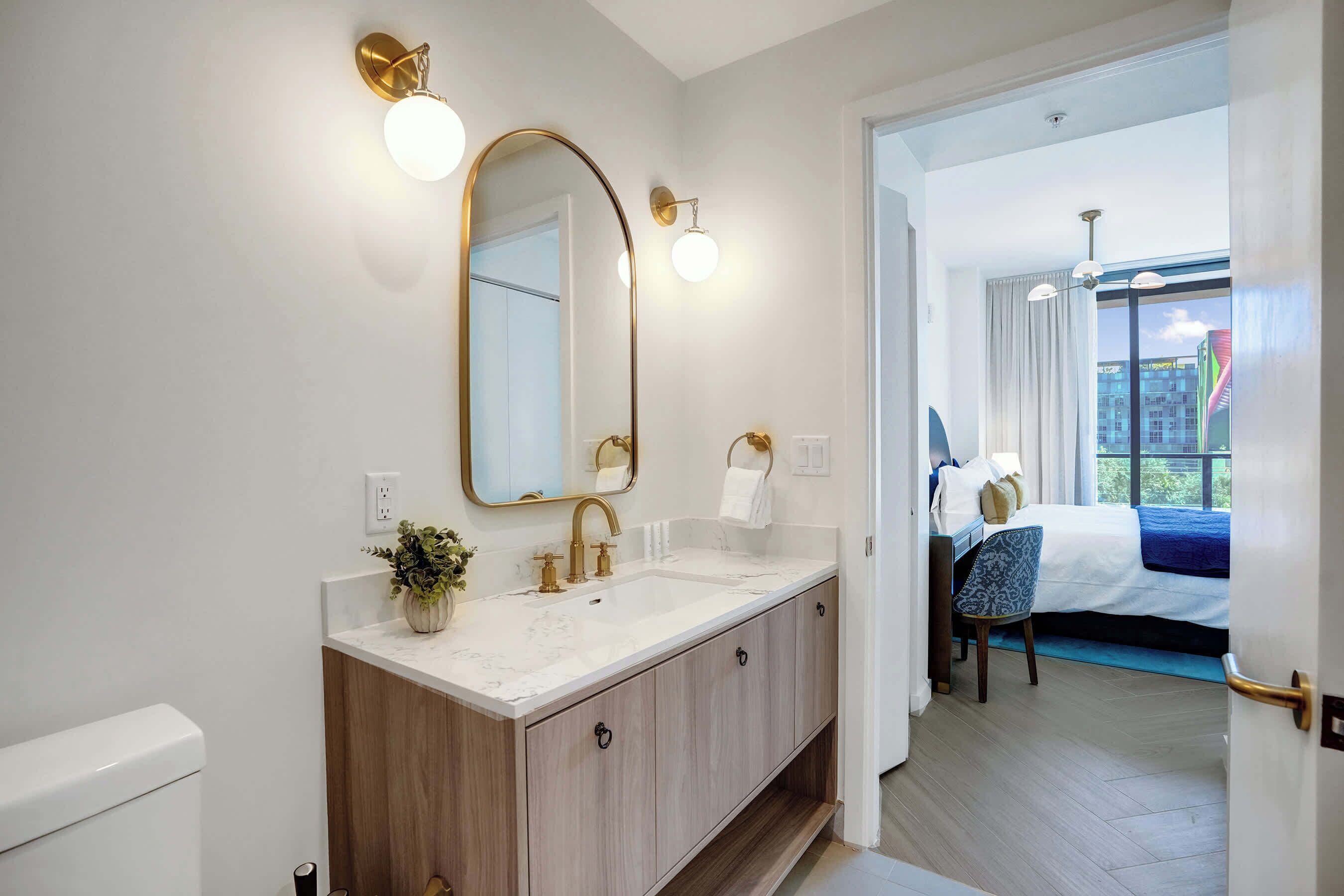 Ein Badezimmer mit einem großen Spiegel, einem Waschbecken und einer Toilette. Eine Tür führt in ein Schlafzimmer mit einem Bett und einem Fenster.