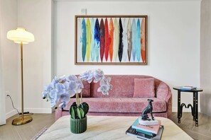 Un soggiorno con un divano rosa, una lampada da terra, un tavolo con libri e una pianta, e un quadro astratto colorato appeso alla parete.