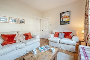 A living room with two white sofas, red and plaid cushions, a wooden coffee table with a magazine and a mug, and a framed picture on the wall. - 3 Bed in Keswick (oc-sz542) (Keswick)