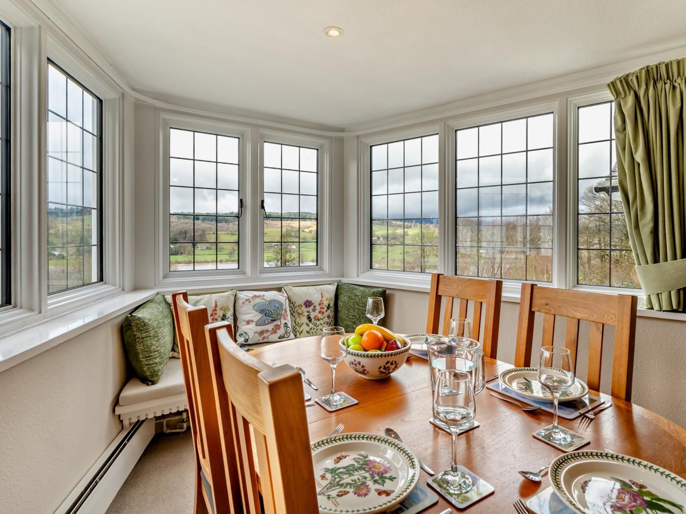 A dining room with a table set for two, a bowl of fruit, and a view of the countryside through large windows.