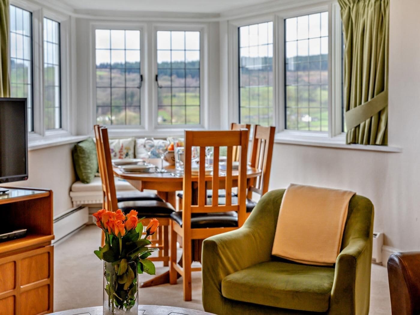 A dining area with a table set for four, surrounded by wooden chairs, a television, and a vase with flowers.
