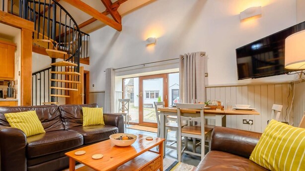 A cozy living room with a brown leather sofa, a wooden coffee table, a spiral staircase, and a TV mounted on the wall. - 3 Bed in Cleator (oc-88627) (Ennerdale and Kinniside)