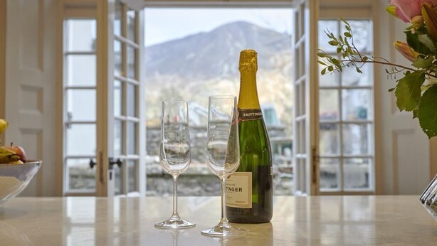 Una botella de champán Taittinger, dos copas y un cuenco de fruta sobre una mesa con vistas a montañas a través de amplios ventanales.