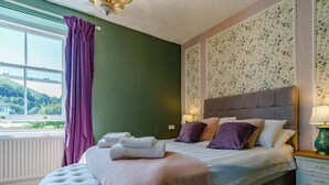 Un dormitorio con una cama grande, un otoman tapizado, una ventana con cortinas y un papel tapiz floral.