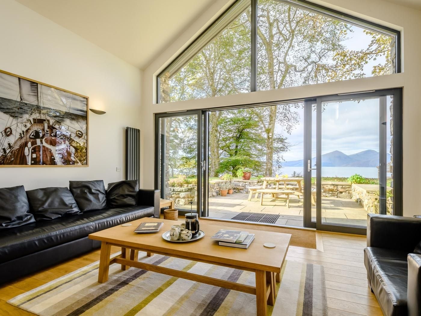 A spacious living room with a large window, a black leather sofa, a wooden coffee table, and a painting on the wall.