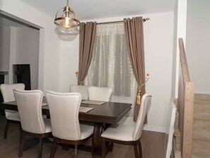 A dining area with a wooden table, six upholstered chairs, a chandelier, and a window with curtains.