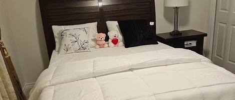 Uma cama bem arrumada com edredom branco e almofadas, uma luminária de cabeceira e um criado-mudo pequeno.