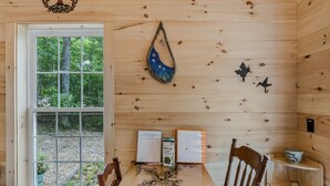 Een houten cabin interieur met een eettafel, stoelen, een raam met uitzicht op bomen, een decoratie en een klein boekenrek.